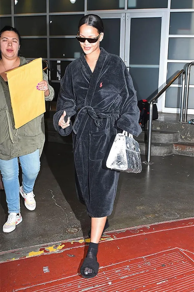 A person wearing sunglasses, a black robe, and slipper sandals carries a silver handbag, accompanied by another person.