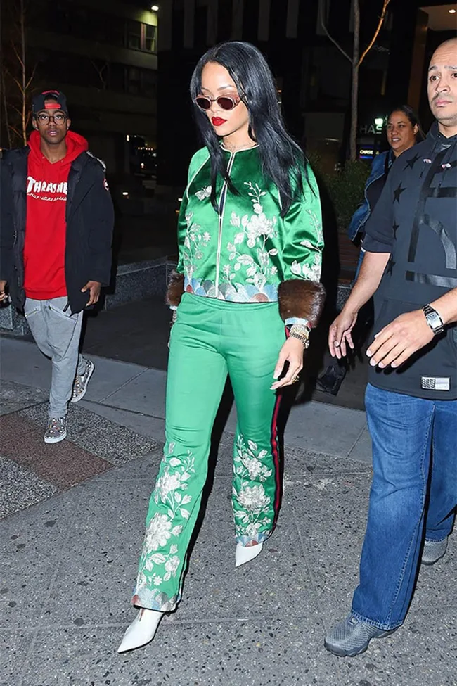 Woman in a green floral outfit and sunglasses walking on a city street at night, surrounded by others.