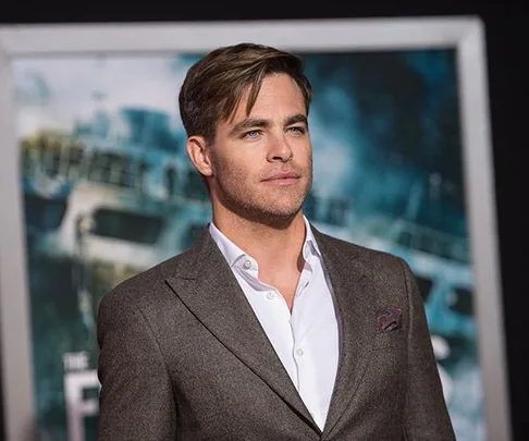 A man in a brown suit with a white shirt at a movie premiere, with a ship on the background poster.