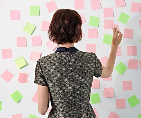 Person facing wall with pink and green sticky notes, wearing a patterned dress, reaching to place or remove a note.