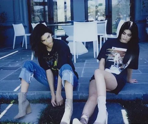 Two women sitting casually outdoors near a patio, wearing graphic t-shirts and ankle boots.