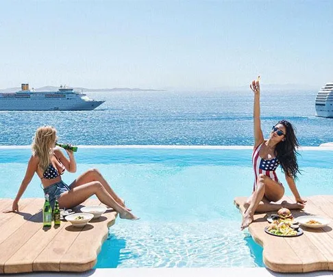 Two women in swimsuits by an infinity pool, with ocean liners in the background; one raises her arm, celebrating.