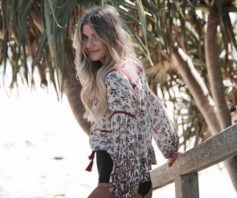 Woman with long blonde hair in a floral blouse standing by a fence, palm trees in the background at Byron Bay.