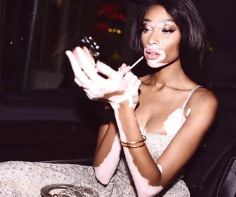 Model applying makeup in car, wearing a sleeveless outfit with gold accessories. She has distinctive skin markings.