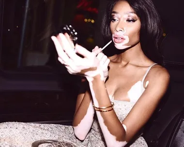 Model applying makeup in car, wearing a sleeveless outfit with gold accessories. She has distinctive skin markings.