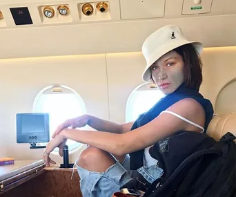 Woman in a white hat and casual attire sitting on an airplane seat with a green face mask on her face.