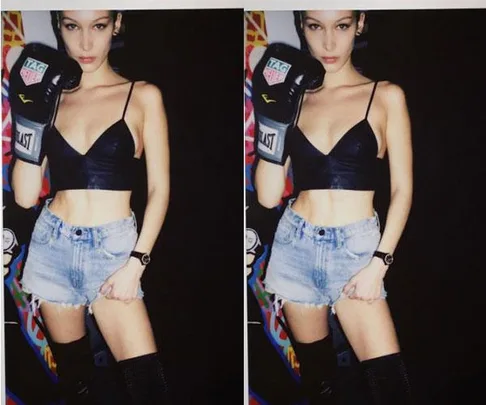 Bella Hadid posing in black crop top, denim shorts, and boxing gloves, standing in front of a colorful background.