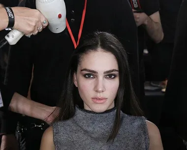 A woman with long hair getting a blow-dry by a stylist backstage at a fashion show.