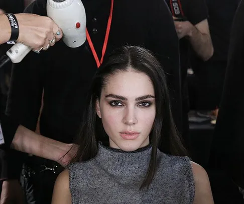 A woman with long hair getting a blow-dry by a stylist backstage at a fashion show.