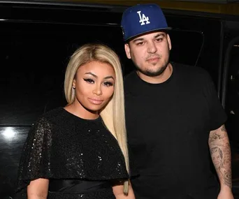 Blac Chyna and Rob Kardashian are posing together, Blac Chyna in a black dress, and Rob Kardashian in a black shirt and blue cap.