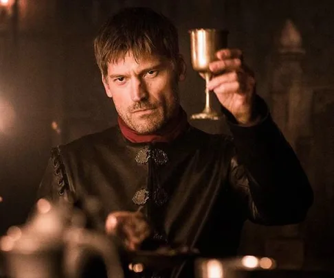 Jaime Lannister, dressed in black, holding up a goblet in a dimly lit room from the Game of Thrones TV show.