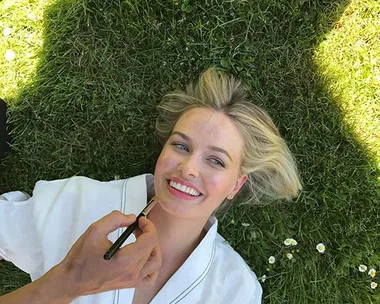 A woman with blonde hair is lying on grass, smiling as someone applies makeup to her face.