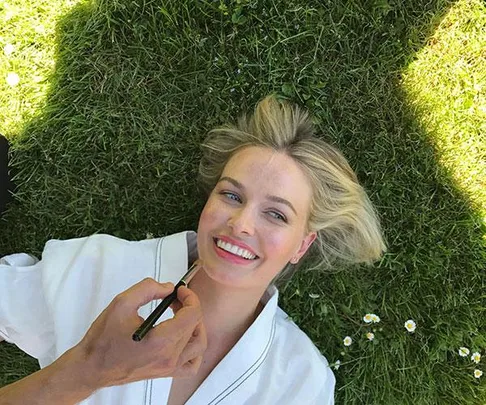 A woman with blonde hair is lying on grass, smiling as someone applies makeup to her face.