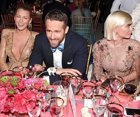 Three seated at a formal event, one laughs, one smiles, one looks away, table set with glasses, flowers, dressed formally.