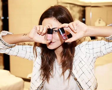 Woman posing with two nail polish bottles covering her eyes, wearing a white tweed jacket and white shirt.