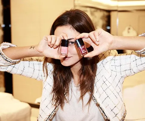 Woman posing with two nail polish bottles covering her eyes, wearing a white tweed jacket and white shirt.