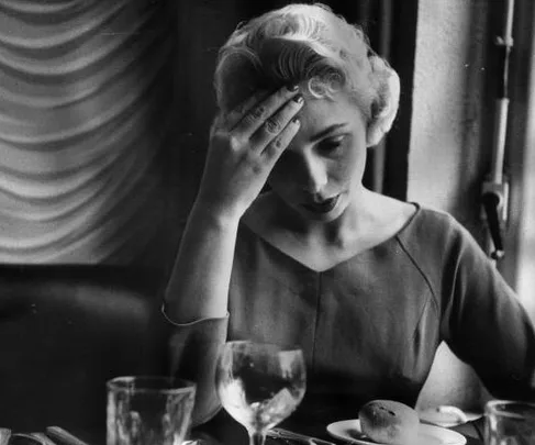 A woman holding her head at a restaurant table, appearing to have a headache, with dining items including glasses and bread in front of her.