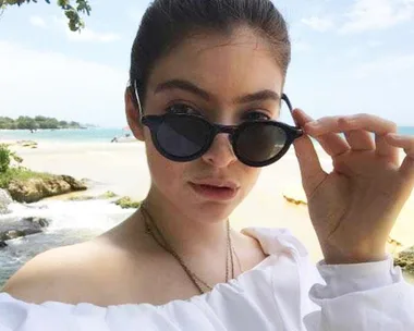 Woman in sunglasses at the beach, adjusting her shades, wearing an off-shoulder white top.