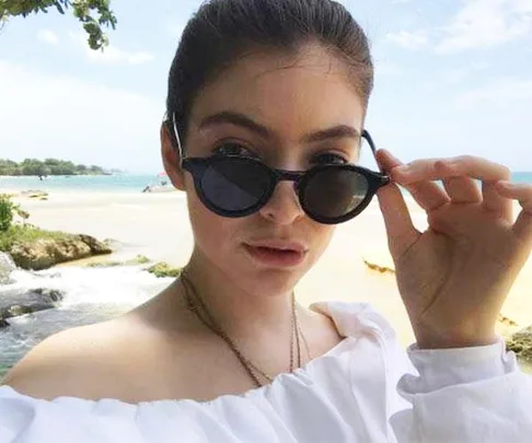 Woman in sunglasses at the beach, adjusting her shades, wearing an off-shoulder white top.