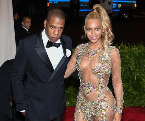 A man in a black tuxedo and a woman in a sheer, jeweled gown pose on a red carpet event.