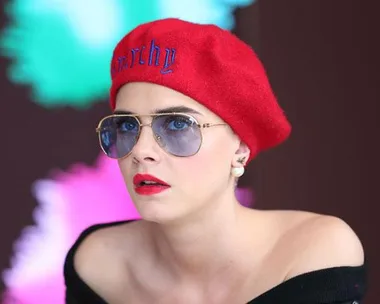 Woman with short hair in red beret and sunglasses, wearing off-shoulder top with colorful background.