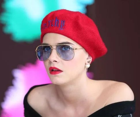 Woman with short hair in red beret and sunglasses, wearing off-shoulder top with colorful background.