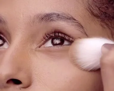 A close-up of an eye, with a makeup brush applying translucent powder to the face.