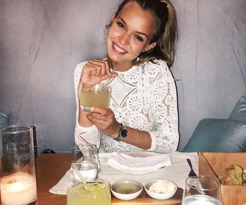 Woman in a white lace top smiling and holding a drink at a restaurant table with candles and appetizers.