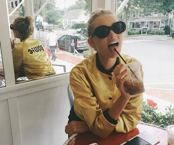 Woman in a gold jacket and black sunglasses drinks iced coffee at an outdoor cafe with a window reflection behind her.