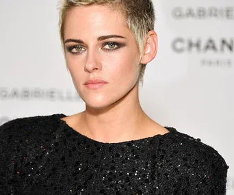 Kristen Stewart with short blonde hair wearing a black sequin top at an event, posing against a white background.