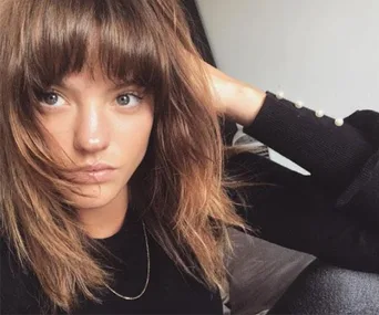 A close-up of a woman with light brown hair and bangs, wearing a black top, resting her arm on her head in a casual pose.