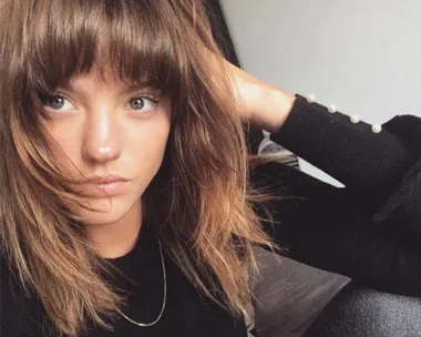 A close-up of a woman with light brown hair and bangs, wearing a black top, resting her arm on her head in a casual pose.