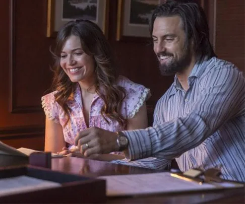 Two characters from "This Is Us," Rebecca and Jack, smile while looking at a document in a warmly lit room.