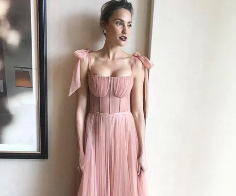 Woman in a blush pink gown with bow straps, standing indoors by a framed picture.