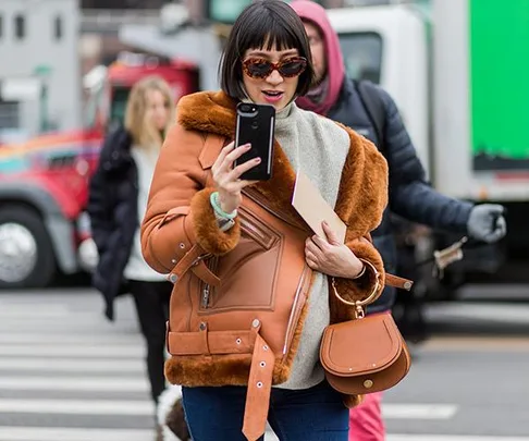 A stylish woman with sunglasses and a brown fur-lined jacket takes a selfie on a city street.