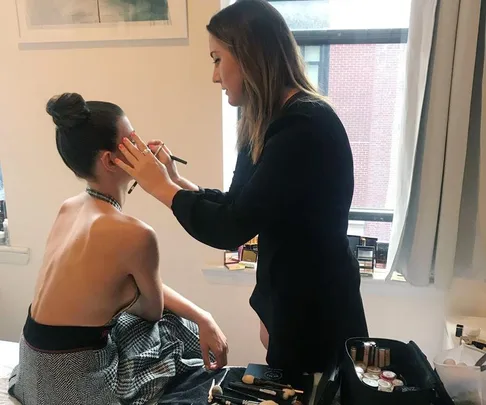 A makeup artist applies eyeshadow to a woman with a backless dress seated near a window, with makeup tools spread out beside them.