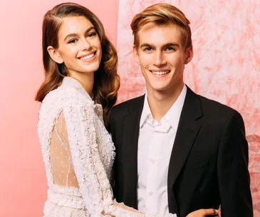 Two people smiling, one in a white dress and the other in a black suit, against a pink background.