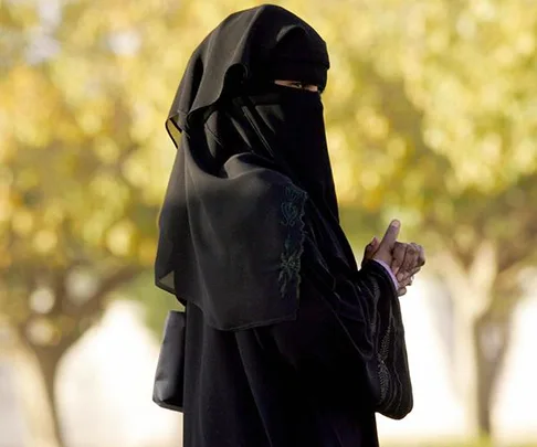 A woman in a black niqab stands outdoors, with blurred trees in the background.