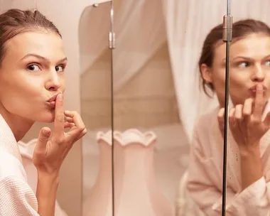 Woman in bathrobe looking in bathroom mirror, touching her lips with finger.