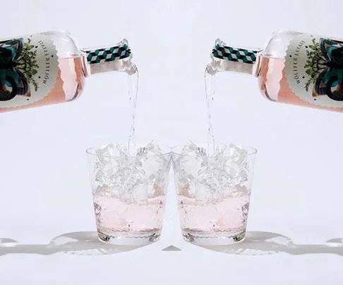 Two bottles of pink gin being poured into glasses filled with ice against a white background.