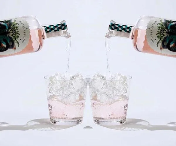 Two bottles of pink gin being poured into glasses filled with ice against a white background.