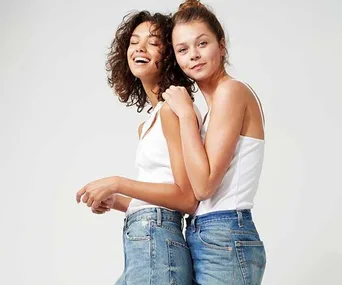 Two women wearing white tops and denim jeans, smiling, and posing against a plain background.