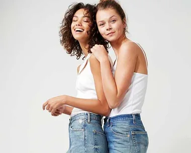 Two women wearing white tops and denim jeans, smiling, and posing against a plain background.