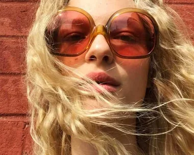 Woman with long curly blonde hair wearing large orange sunglasses against a brick wall.