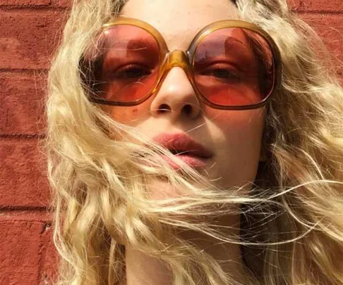 Woman with long curly blonde hair wearing large orange sunglasses against a brick wall.