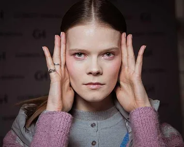 Young woman with hands on the sides of her face, looking serious and wearing a grey top and pink sweater.