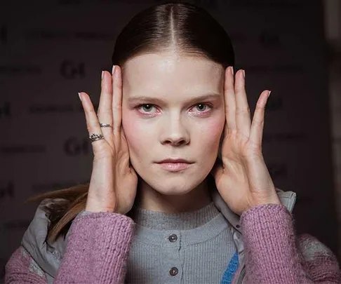 Young woman with hands on the sides of her face, looking serious and wearing a grey top and pink sweater.