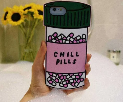 Hand holding phone case resembling pill bottle labeled "CHILL PILLS" in front of a bathtub with flowers in background.