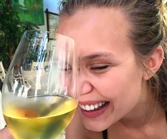 A woman smiling while holding a large glass of white wine close to her face.