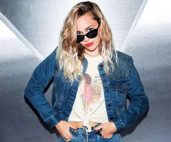 Miley Cyrus in denim jacket and sunglasses, posing with hands on hips in front of a metallic background.